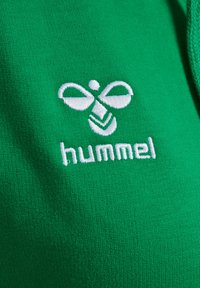 Grüner Baumwollstoff mit einem weißen gestickten Logo, das stilisierte Bienelemente zeigt, und dem Text "hummel" darunter.