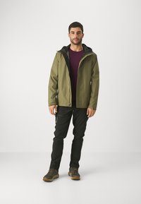 Giacca verde oliva impermeabile con cappuccio nero, abbinata a una camicia bordeaux e pantaloni neri. Materiale resistente e design pratico.