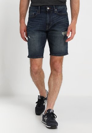 Adulte portant un short en denim bleu foncé usé et des baskets noires New Balance, marchant sur un fond blanc uni.