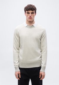 Jeune homme aux cheveux bouclés portant un pull en maille beige par-dessus une chemise blanche à col et un pantalon noir, debout devant un fond clair uni.