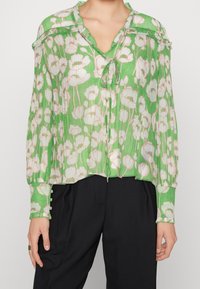 Groene blouse met bloemenpatroon van doorschijnende, geplooide stof. Heeft een V-hals, knoopmanschetten en een strikdetail bij de halslijn.