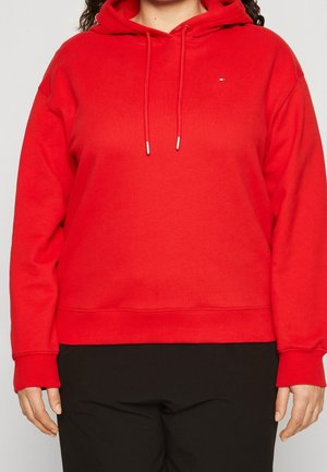 Personne portant un sweat à capuche rouge vif avec cordons et un pantalon noir, montrée du cou aux hanches sur un fond uni.
