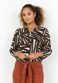 Blusa a maniche lunghe con un motivo geometrico nei colori nero, crema e ruggine. Presenta un scollo a V e una vestibilità comoda. Abbinata a pantaloni color ruggine.