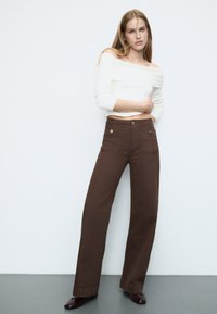 Brown wide-leg pants with two front pockets and metal buttons, paired with a white off-shoulder sweater. Smooth texture and casual design.