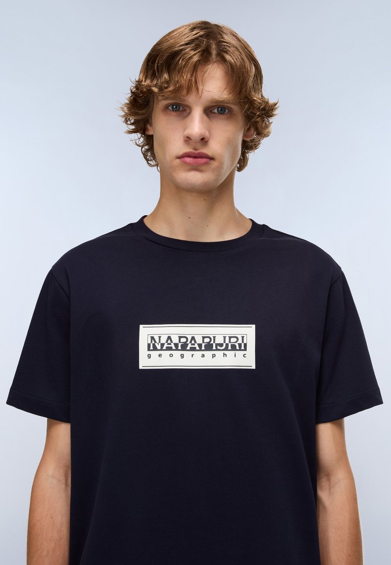 T-shirt in cotone blu navy con una grafica rettangolare bianca con il testo "NAPAPIJRI geographic" in nero, maniche corte e scollatura a girocollo.