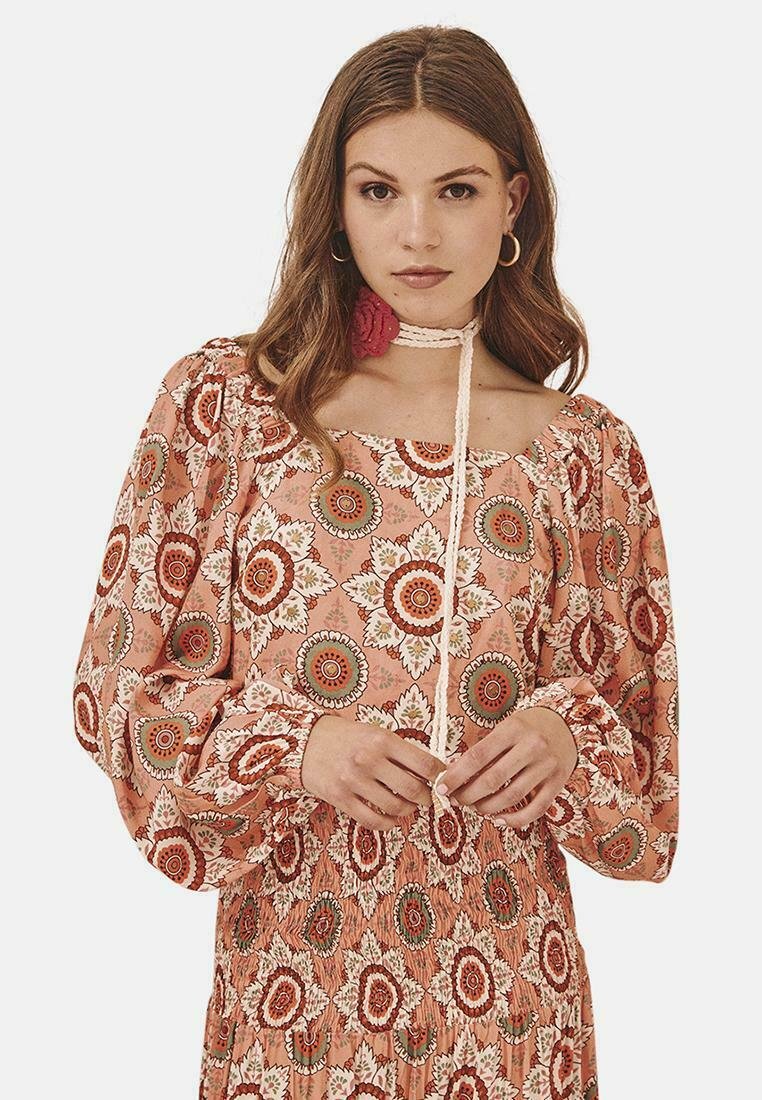 Vestido de manga larga, con cuello descubierto en color coral y estampados florales en rojo, blanco y verde. Presenta una cintura fruncida y un detalle de lazo en el cuello.