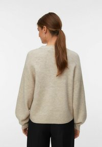 Donna con capelli castani raccolti in una coda bassa, che indossa un maglione beige e pantaloni neri, di spalle contro uno sfondo chiaro e uniforme.