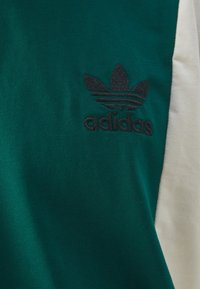 Grünes Baumwoll-T-Shirt mit einem schwarzen Adidas-Logo, das auf der linken Brust eingestickt ist, und einem cremefarbenen Ärmelakzent. Weiche Textur, lässiges Design.
