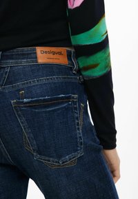 Tamsiai mėlynos džinsinės kelnės su tekstūruotu paviršiumi, turi dvi galines kišenes ir rudą odinę etikete su užrašu "Desigual. PREMIUM DENIM."