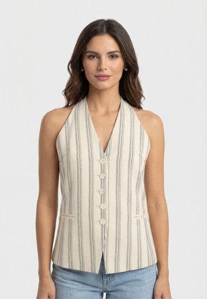 Mujer con chaleco beige sin mangas con rayas negras verticales, abotonado al frente, combinado con vaqueros azul claro, de pie contra un fondo liso.