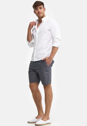 Camicia bianca di lino con bottoni, pantaloncini grigi e scarpe bianche slip-on. La camicia ha una vestibilità comoda, maniche lunghe e un colletto. I pantaloncini arrivano al ginocchio.