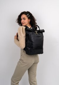 Black leather backpack with a roll-top design, gold hardware, and side pocket. Smooth texture with stitching details.