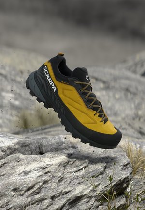 Kollane ja must Scarpa Gore-Tex matkajalats õhus kivise maastiku kohal, näidates vastupidavat tallat ja paelu.