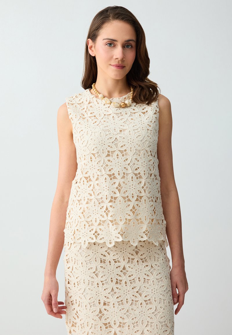 Femme portant un top en dentelle crème sans manches et une jupe assortie à motif floral, accessoirisée d'un collier épais en perles, sur fond uni.