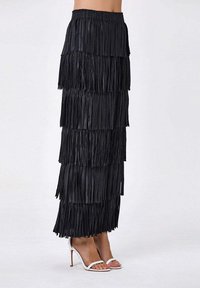 Jupe longue noire à franges avec des couches superposées et une taille élastique. Associée à des talons blancs à bouts ouverts pour un contraste.