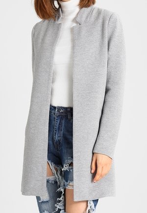 Femme portant un long cardigan texturé gris clair sur un pull à col roulé blanc et un short en jean bleu déchiré.