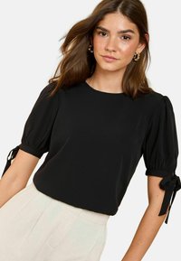 Blouse noire à manches bouffantes avec des accents noués aux poignets, texture lisse et encolure ronde. Associée à un pantalon de couleur claire.