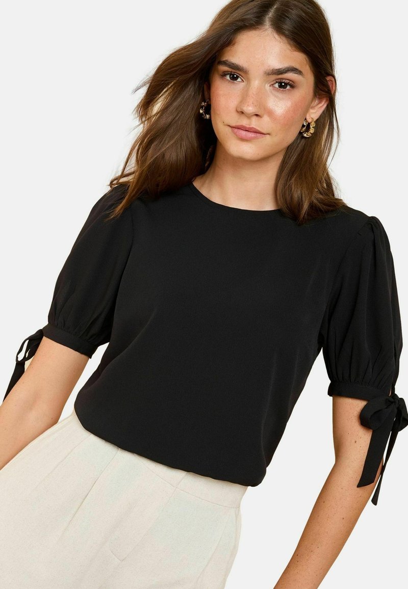 Blouse noire à manches bouffantes avec des accents noués aux poignets, texture lisse et encolure ronde. Associée à un pantalon de couleur claire.