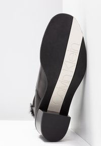 Zapato de cuero negro con superficie texturizada, que presenta un tacón cuadrado y un prominente logo blanco de Calvin Klein en la suela.