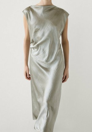 Femme portant une robe midi sans manches en satin argenté avec un col bateau et une texture légèrement froissée, debout devant un fond uni.