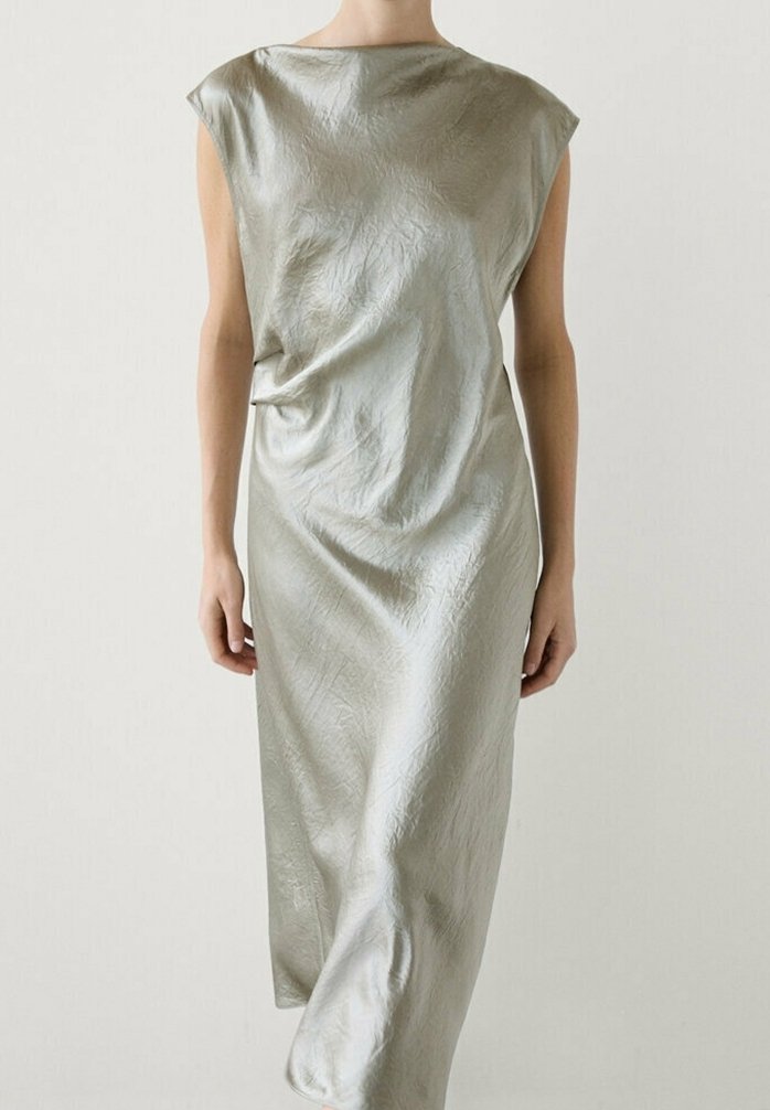 Femme portant une robe midi sans manches en satin argenté avec un col bateau et une texture légèrement froissée, debout devant un fond uni.