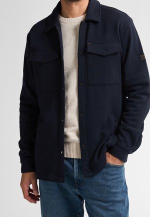 Mand iført navyblå knappet jakke over beige sweater og blå jeans, den ene hånd i lommen mod ensfarvet baggrund.