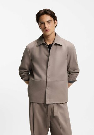 Ung mand med kort mørkt hår iført en taupe knappet jakke over matchende løse bukser med stofbælte, stående med hænderne bag ryggen.