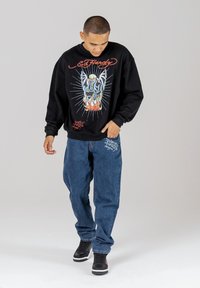 Black sweatshirt with graphic design featuring flames and a skeleton, paired with loose blue jeans. Black sneakers with white and red accents.