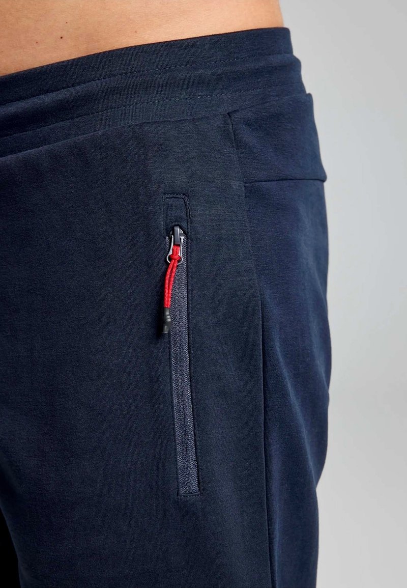 Section de pantalon bleu marine avec une poche zippée dotée de détails en rouge. Le tissu a une texture lisse, conçu pour le confort et l'utilité.