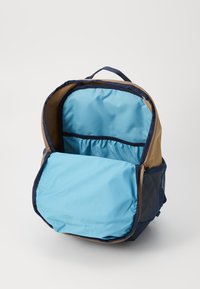 Ryggsekk med en lys brun ytre, marineblå detaljer og blå innvendig. Har en frontlomme og en nettinglomme på siden. Glidelåsåpning for enkel tilgang.
