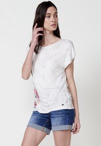 Chemise blanche à motif floral avec une coupe ample, un col rond et des manches courtes. Associée à un short en denim bleu avec des ourlets retroussés.
