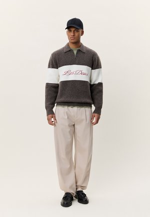 Καφέ πλεκτό πουλόβερ με λευκή οριζόντια ρίγα και κόκκινο κείμενο «Les Deux», συνδυασμένο με μπεζ παντελόνια και μαύρα παπούτσια. Μαύρο απλό καπέλο.