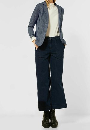 Femme portant un blazer texturé bleu, un haut blanc à col montant, un pantalon en jean large et foncé, et des bottines à lacets noires, les mains dans les poches.