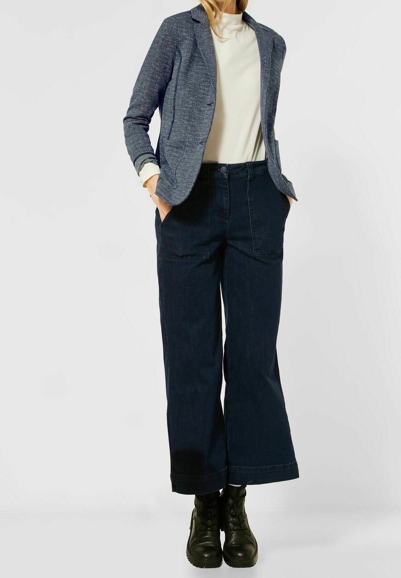 Donna che indossa un blazer blu strutturato, un top bianco a collo alto, jeans scuri a gamba larga e stivaletti neri con lacci, con le mani in tasca.