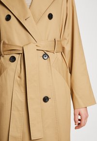 Abrigo trench beige con diseño de doble botonadura, botones negros, cinturón atado y bolsillos laterales, que muestra una tela suave y texturizada.