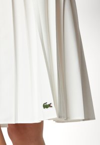 White pleated skirt made from smooth fabric with an embroidered green crocodile logo at the hem. Features a layered design texture.