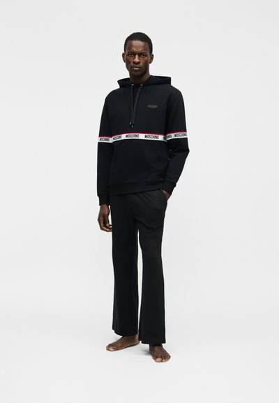 Άνδρας που στέκεται ξυπόλητος, φοράει μαύρη κουκούλα με λευκές ταινίες λογότυπου Moschino στο στήθος και τα χέρια, συνδυασμένη με μαύρο παντελόνι φαρδύ στο πόδι.