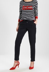 Granatowo-biały sweter w paski z czerwonym napisem „Bonjour Madame”, zestawiony z ciemnymi spodniami i czerwonymi butami na wysokim obcasie. Miękka dzianinowa faktura.