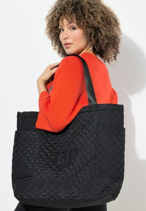 Vrouw met krullend haar die een rode trui draagt en een grote zwarte gewatteerde draagtas met zwarte bandjes over haar schouder draagt.