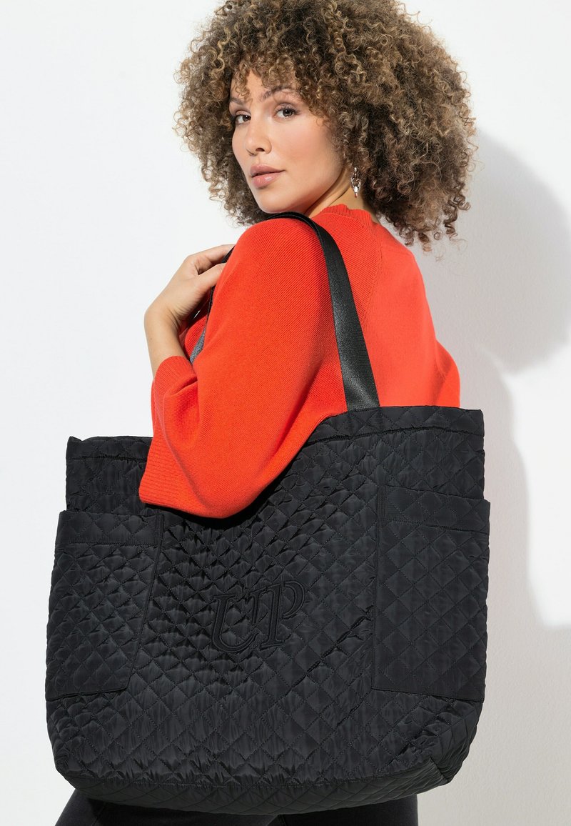 Frau mit lockigem Haar, die einen roten Pullover trägt und eine große schwarze gesteppte Tragetasche mit schwarzen Trägern über der Schulter trägt.