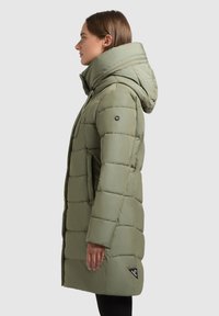 Olive zaļas pufkoats ar kapuci, ar iešuvēm, sānu kabatām un logotipa uzkodi apakšējā kreisajā pusē.
