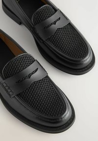 Zwarte loafers hebben een geweven textuur op de bovenkant, gladde leren accenten en een contrasterende rubberen zool. Geen zichtbare logo's of patronen.