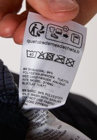 Wasinstructielabel van witte stof met zwarte tekst, pictogrammen voor wasinstructies en materiaaldetails: 94% polyester, 6% spandex.