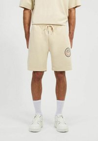 Pantalones cortos de sudadera beige con cinturilla elástica, detalles de cordón y un parche de logo circular en el muslo izquierdo. Textura suave.