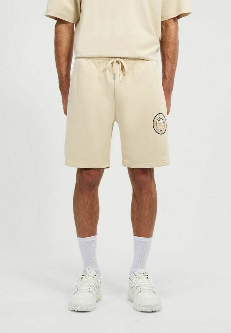 Pantalones cortos de sudadera beige con cinturilla elástica, detalles de cordón y un parche de logo circular en el muslo izquierdo. Textura suave.