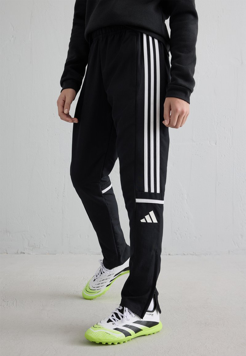 Person wearing black Adidas track pants with white stripes and white-green soccer shoes standing against a gray wall.
