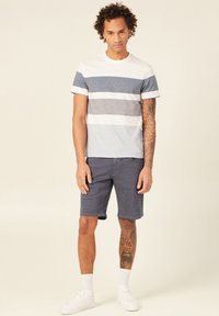 Raidoitu t-paita valkoisena, vaaleansinisenä ja tummansinisenä yhdistettynä harmaisiin shortsit. Asusteena valkoiset tennarit ja valkoiset nilkkasukat.