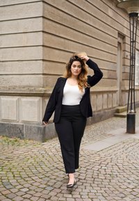 Blazer nero su una maglietta bianca, abbinato a pantaloni neri e tacchi neri. Tessuto liscio, design strutturato, mentre cammina lungo un sentiero lastricato.