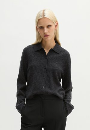 Vrouw met steil blond haar, gekleed in een donkergrijze gebuttonde trui en zwarte broek, staand met handen in de zakken tegen een effen achtergrond.