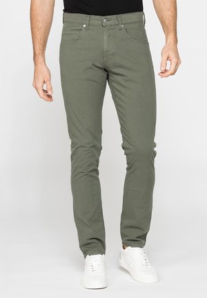 Uomo che indossa pantaloni slim fit verde oliva e sneakers bianche, in piedi davanti a uno sfondo chiaro e uniforme.